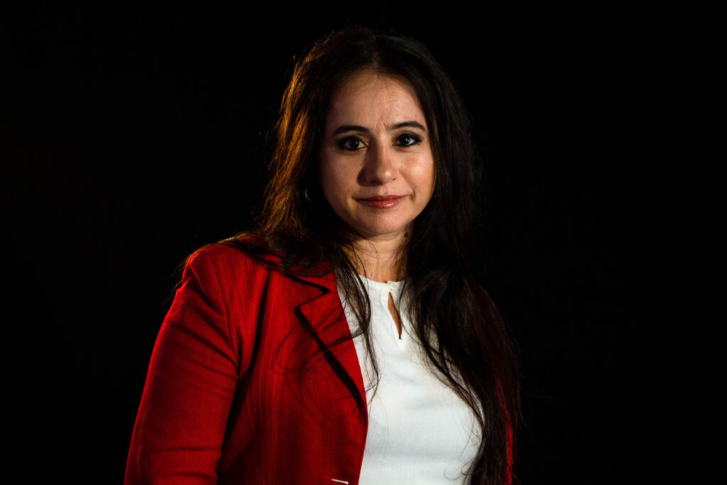 Laura Utrera looks directly at the camera and wears a white shirt and red jacket. The photo background is black.